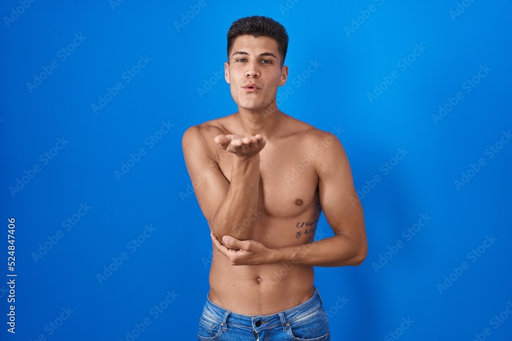Young hispanic man standing shirtless over blue background looking at the camera blowing a kiss with hand on air being lovely and sexy. love expression.