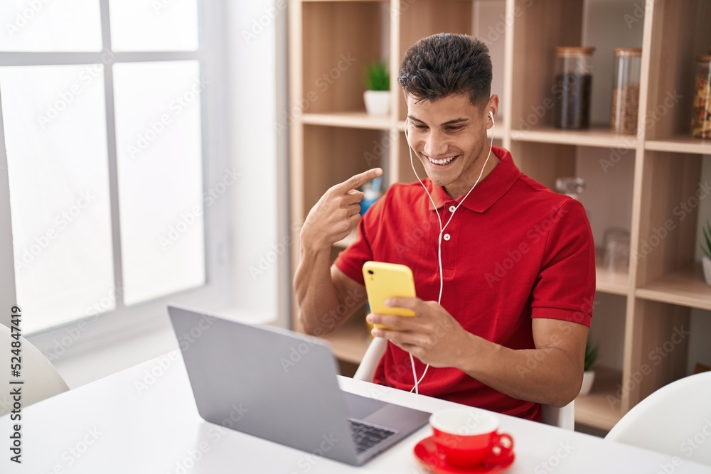 Young hispanic man using laptop and doing video call with smartphone smiling happy pointing with hand and finger Young hispanic man using laptop and doing video call with smartphone smiling happy pointing with hand and finger
