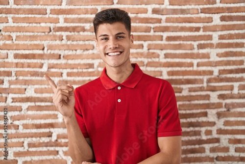 Young hispanic man standing over bricks wall with a big smile on face, pointing with hand and finger to the side looking at the camera.