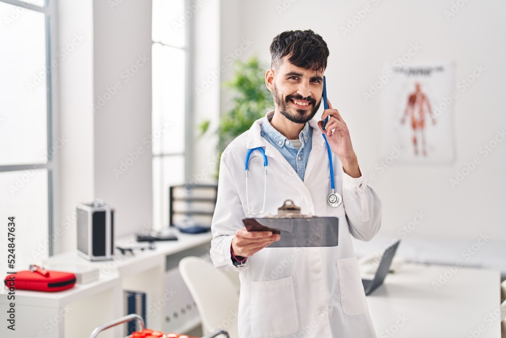 Young hispanic man wearing doctor uniform talking on smartphone reading medical report at clinic Young hispanic man wearing doctor uniform talking on smartphone reading medical report at clinic