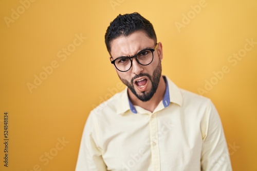 Hispanic young man wearing business clothes and glasses in shock face, looking skeptical and sarcastic, surprised with open mouth