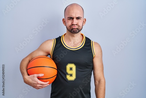 Young bald man with beard wearing basketball uniform holding ball puffing cheeks with funny face. mouth inflated with air, crazy expression.