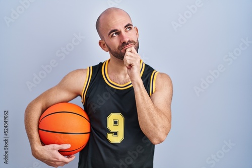 Young bald man with beard wearing basketball uniform holding ball with hand on chin thinking about question, pensive expression. smiling with thoughtful face. doubt concept.