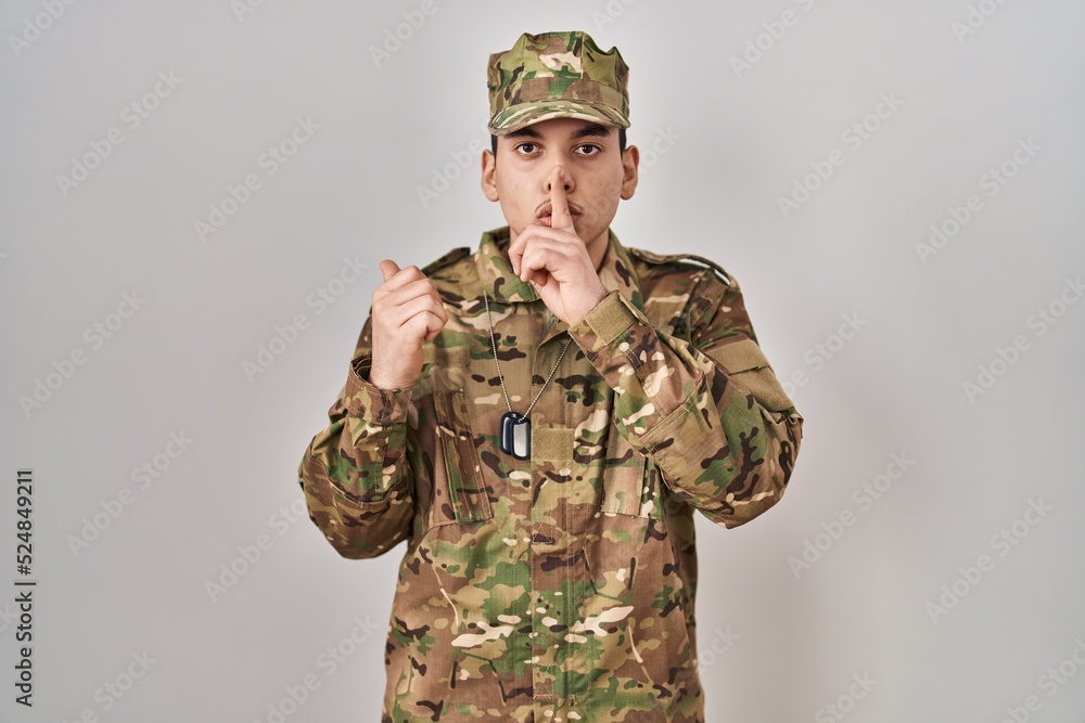 Young arab man wearing camouflage army uniform asking to be quiet with finger on lips pointing with hand to the side. silence and secret concept. Young arab man wearing camouflage army uniform asking to be quiet with finger on lips pointing with hand to the side. silence and secret concept.