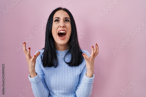Hispanic woman standing over pink background crazy and mad shouting and yelling with aggressive expression and arms raised. frustration concept.