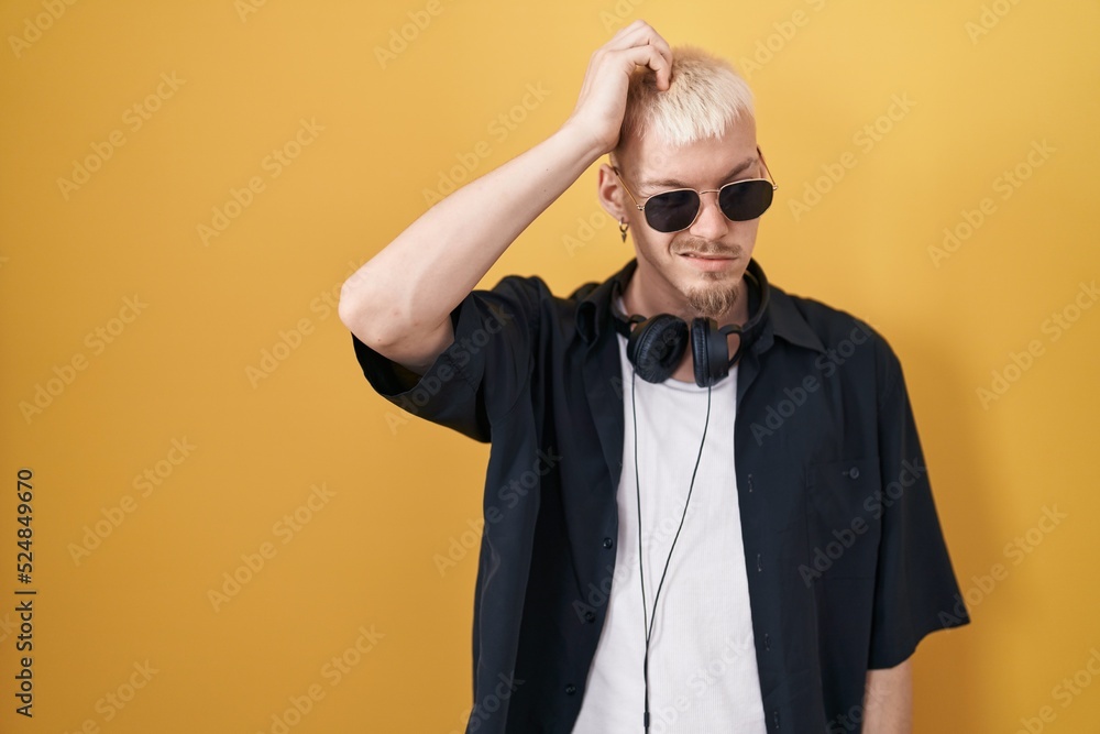 Young caucasian man wearing sunglasses standing over yellow background confuse and wondering about question. uncertain with doubt, thinking with hand on head. pensive concept.