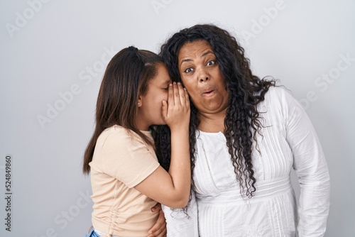 Mother and young daughter standing over white background hand on mouth telling secret rumor, whispering malicious talk conversation