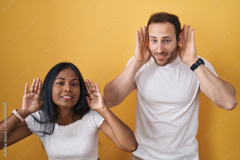 Interracial couple standing over yellow background trying to hear both hands on ear gesture, curious for gossip. hearing problem, deaf Interracial couple standing over yellow background trying to hear both hands on ear gesture, curious for gossip. hearing problem, deaf