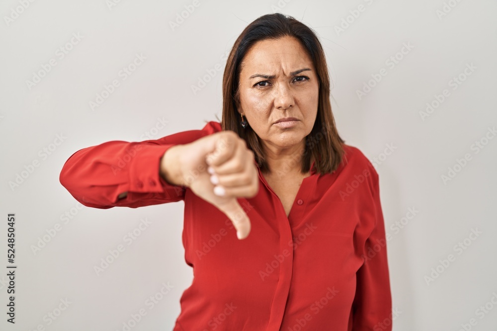 Hispanic mature woman standing over white background looking unhappy and angry showing rejection and negative with thumbs down gesture. bad expression. Hispanic mature woman standing over white background looking unhappy and angry showing rejection and negative with thumbs down gesture. bad expression.