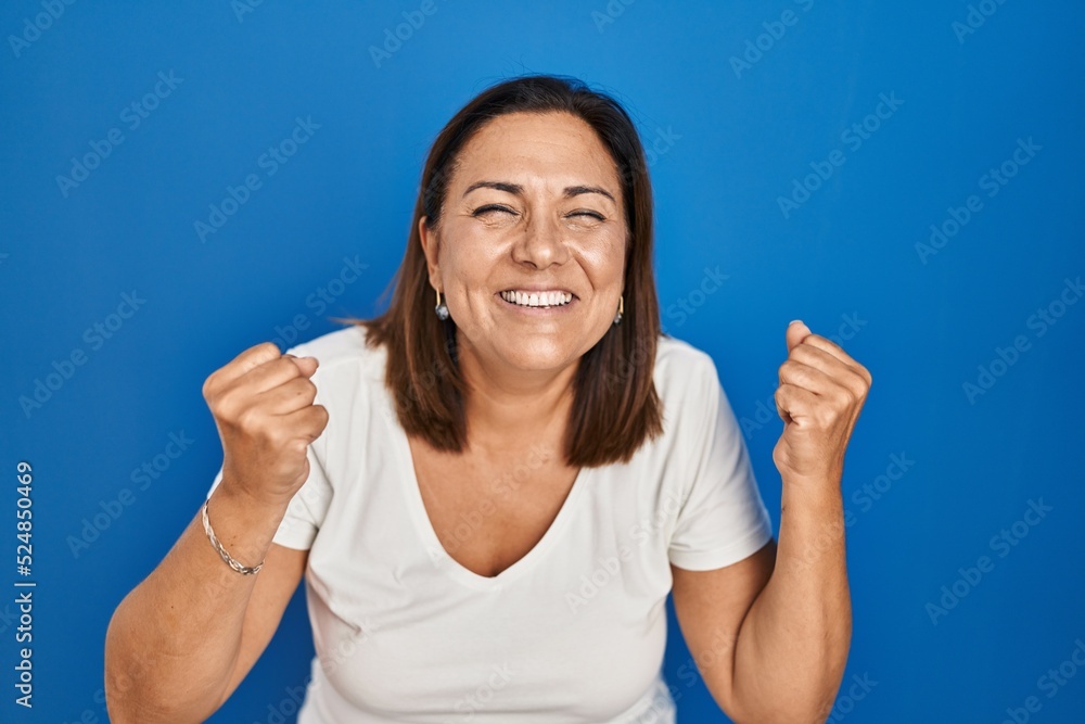 Hispanic mature woman standing over blue background very happy and excited doing winner gesture with arms raised, smiling and screaming for success. celebration concept. Hispanic mature woman standing over blue background very happy and excited doing winner gesture with arms raised, smiling and screaming for success. celebration concept.