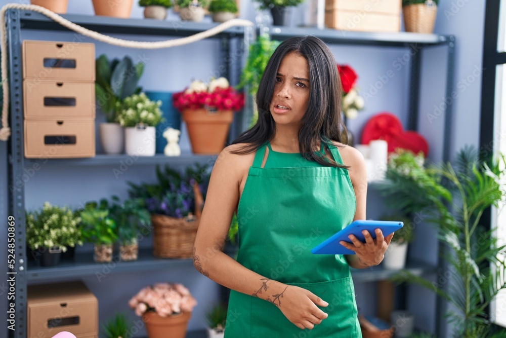 Brunette woman working at florist shop holding tablet in shock face, looking skeptical and sarcastic, surprised with open mouth Brunette woman working at florist shop holding tablet in shock face, looking skeptical and sarcastic, surprised with open mouth