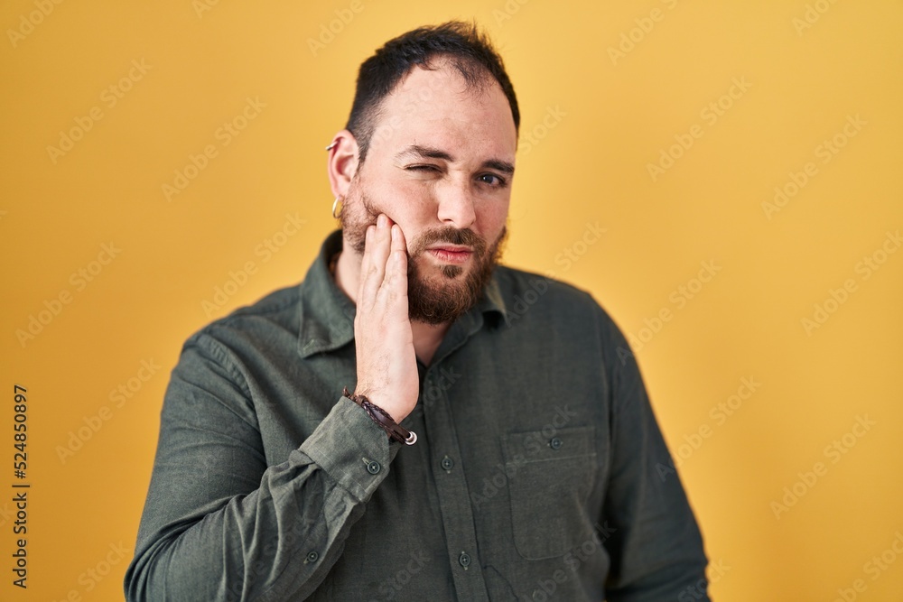 Plus size hispanic man with beard standing over yellow background touching mouth with hand with painful expression because of toothache or dental illness on teeth. dentist Plus size hispanic man with beard standing over yellow background touching mouth with hand with painful expression because of toothache or dental illness on teeth. dentist