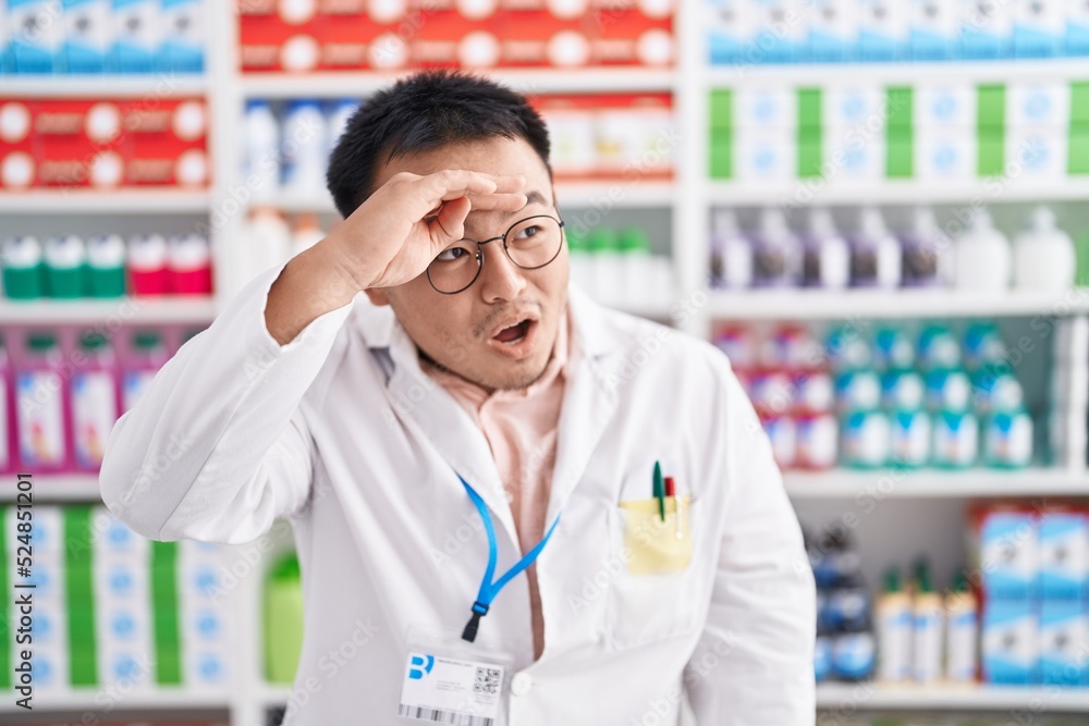 Chinese young man working at pharmacy drugstore very happy and smiling looking far away with hand over head. searching concept. Chinese young man working at pharmacy drugstore very happy and smiling looking far away with hand over head. searching concept.