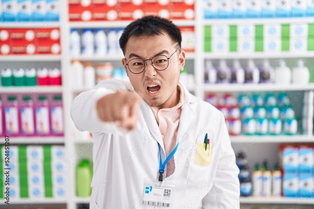 Chinese young man working at pharmacy drugstore pointing displeased and frustrated to the camera, angry and furious with you Chinese young man working at pharmacy drugstore pointing displeased and frustrated to the camera, angry and furious with you