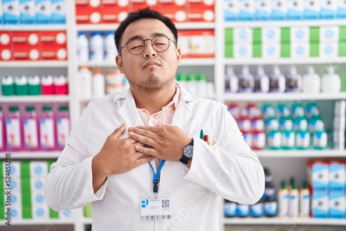 Chinese young man working at pharmacy drugstore smiling with hands on chest with closed eyes and grateful gesture on face. health concept. © Krakenimages.com
