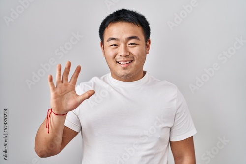 Young chinese man standing over white background showing and pointing up with fingers number five while smiling confident and happy.