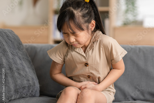 little Asian girl have a stomach ache on couch at home. Child squeeze on her belly so pain and illness. kid has a grimace face. Child health care