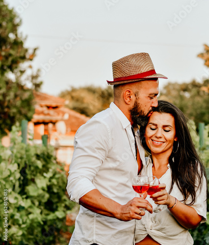 Portrait of a smiling happy couple kissing in a Vineyard toasting wine. Beautiful brunette woman and bearded muscular man spending time together during grape harvest.
