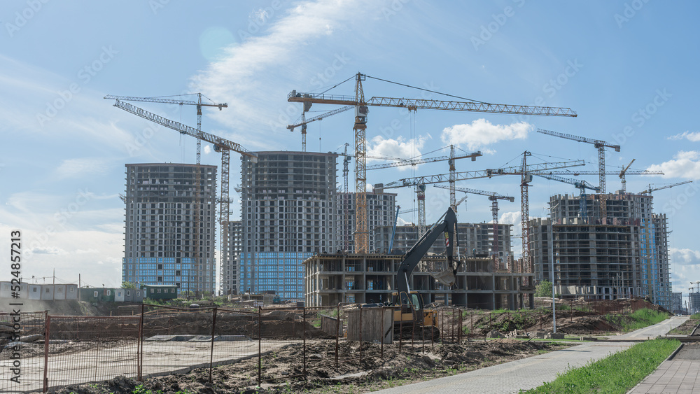 Construction of a multi-story residential buildings. Defocused foreground with young tree. Cranes work. Construction site. Construction of a multi-story residential buildings. Defocused foreground with young tree. Cranes work. Construction site.