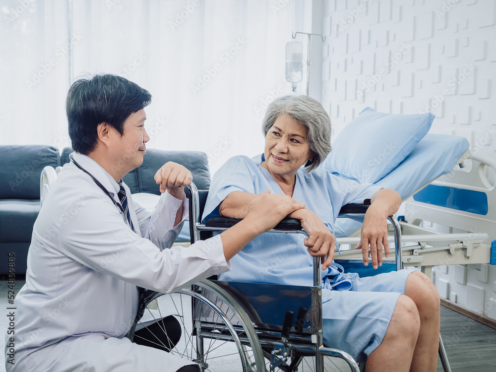 Obraz premium Asian senior woman patient dressed in blue, smiling happily sits in wheelchair with kindly man doctor in white suit holding her hand beside in recovery room in hospital. Healthcare support concept.