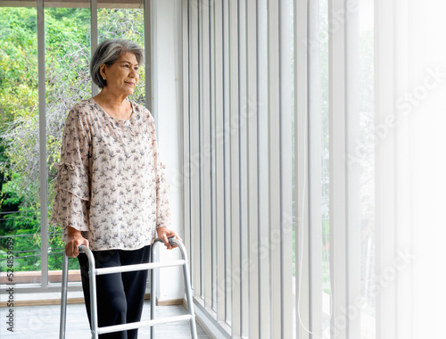 Asian senior woman, white hair standing with walking frame and looking out the glass window, indoors. Elderly lady patient using walking frame. Strong health, medical care and life insurance concepts.