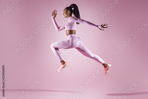 Confident young woman in sports clothing jumping against pink background © gstockstudio
