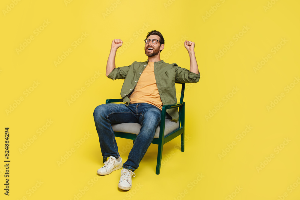 Emotional middle aged man raising fists up, sittign in armchair on yellow studio background, celebrating success Emotional middle aged man raising fists up, sittign in armchair on yellow studio background, celebrating success