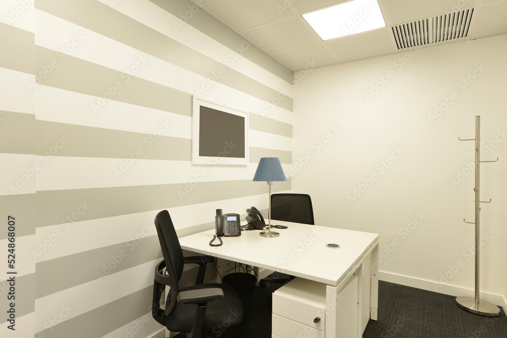 Fototapeta premium Office cubicle with small white double table with telephones, black swivel chairs, metal coat rack, scratched wall and dark gray carpet floor