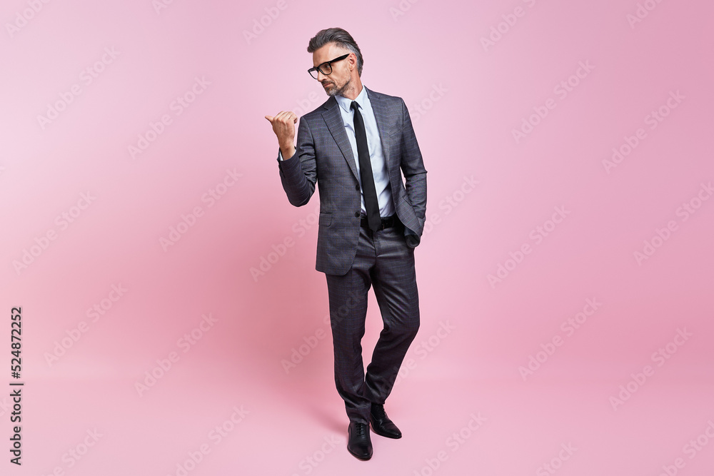 Confident mature man in full suit pointing away while standing against pink background Confident mature man in full suit pointing away while standing against pink background