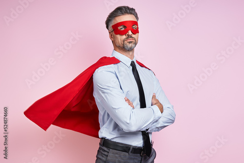 Confident man in shirt and tie wearing superhero cape and keeping arms crossed against pink background photo