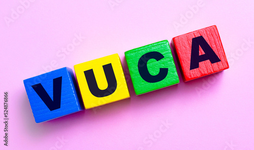 On a light pink background, multi-colored wooden cubes with the text VUCA Volatility Uncertainly Complexity Ambiguity photo