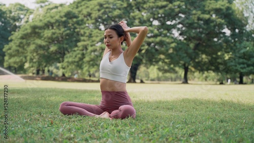 Asian young woman sitting on the grass practicing yoga in the cow pose position in city park with the big trees background. Rearview of female practicing yoga outdoors on a sunny day.