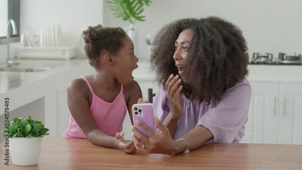 Multi-ethnic young woman with her charming little daughter looking at the camera while using the mobile with surprised expression, watching cartoons, movies, scrolling news feed, relaxing at home