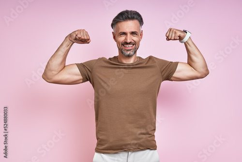Happy mature man showing his biceps while standing against pink background