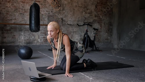 Woman turns on video lesson of training on laptop preparing to repeat after coach. Young blonde beginner lies on black mat to do exercise in gym slow motion photo