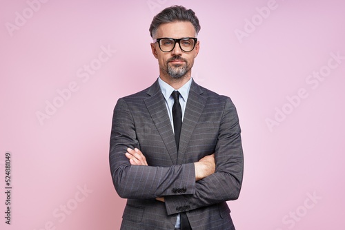 Confident mature man in formalwear keeping arms crossed while standing against pink background