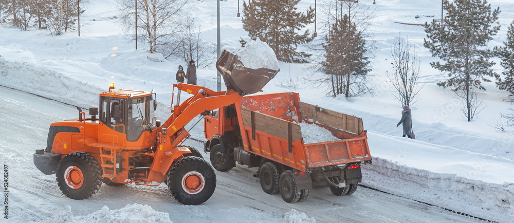 Big orange tractor cleans up snow from the road and loads it into the truck. Cleaning and cleaning of roads in the city from snow in winter. Snow removal after snowfall and blizzards. Big orange tractor cleans up snow from the road and loads it into the truck. Cleaning and cleaning of roads in the city from snow in winter. Snow removal after snowfall and blizzards.