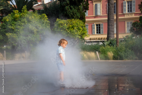 Toddler boy under spray jets of extensive water mirror fountain in Nice, France. Pleasant refreshment , stay coll in summer heat.