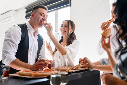 Crop portrait of pretty bride woman feeds the groom with pizza in the kitchen with guests friends of brides. A loving couple celebrating engagement waiting for wedding ceremony.