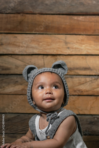 vertical photo of a beautiful latin baby girl with brown skin, wearing overalls and a cap with mouse ears, crocheted with the crochet technique, on a wooden background, with her eyes raised to the sky
