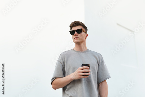 Portrait of handsome confident stylish hipster man in sunglasses holding coffee paper cup, standing guy above white background.