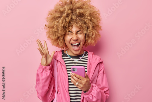 Emotional curly haired woman reacts to awesome news keeps palm raised exclaims loudly holds mobile phone dressed in casual clothing browses internet isolated over pink background. Human reactions