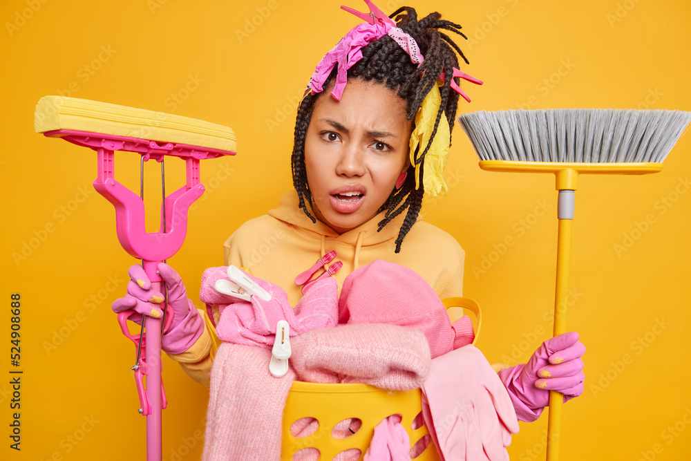 Puzzled confused woman with braided hair holds cleaning mop and brush