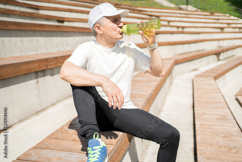 A retired man drinks water from a bottle, sports fitness does a workout in the park.