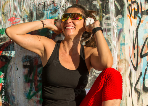 Beautiful smiling young girl listening to music with headphones while resting from doing daily exercise. Sportswoman With running glasses and headphones With a background of a graffiti. Running woman © Nanci
