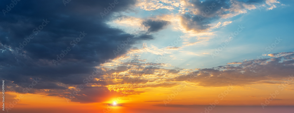 Setting sun in the orange blue evening sky. Sun rays break through the light clouds at sunset. Dark stormy cloud covering half the sky at sundown. Picturesque skyscape wide panorama.