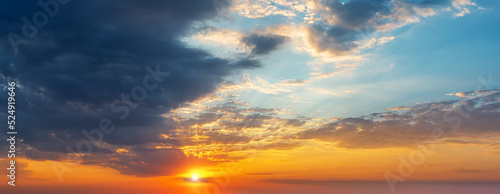 Setting sun in the orange blue evening sky. Sun rays break through the light clouds at sunset. Dark stormy cloud covering half the sky at sundown. Picturesque skyscape wide panorama. © Maryia