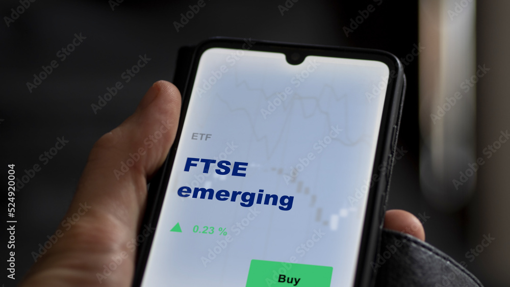An investor's analyzing the FTSE emerging etf fund on screen. A phone shows the ETF's prices London England london ftse england to invest An investor's analyzing the FTSE emerging etf fund on screen. A phone shows the ETF's prices London England london ftse england to invest
