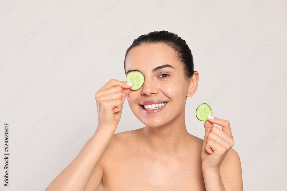 Beautiful young woman putting slices of cucumber on eyes against light grey background Beautiful young woman putting slices of cucumber on eyes against light grey background