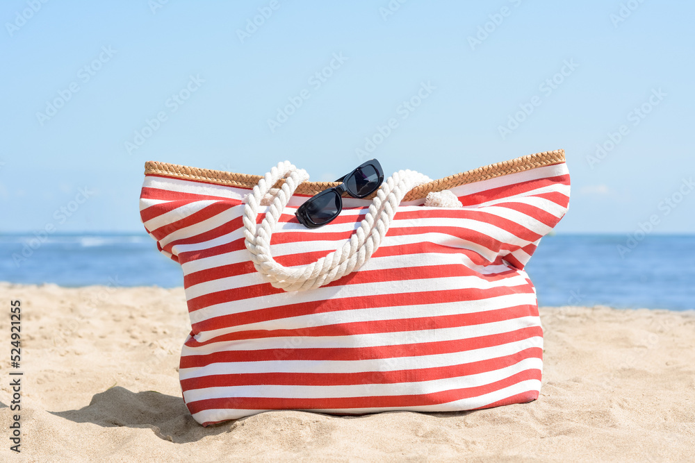 Stylish striped bag with sunglasses on sandy beach near sea Stylish striped bag with sunglasses on sandy beach near sea